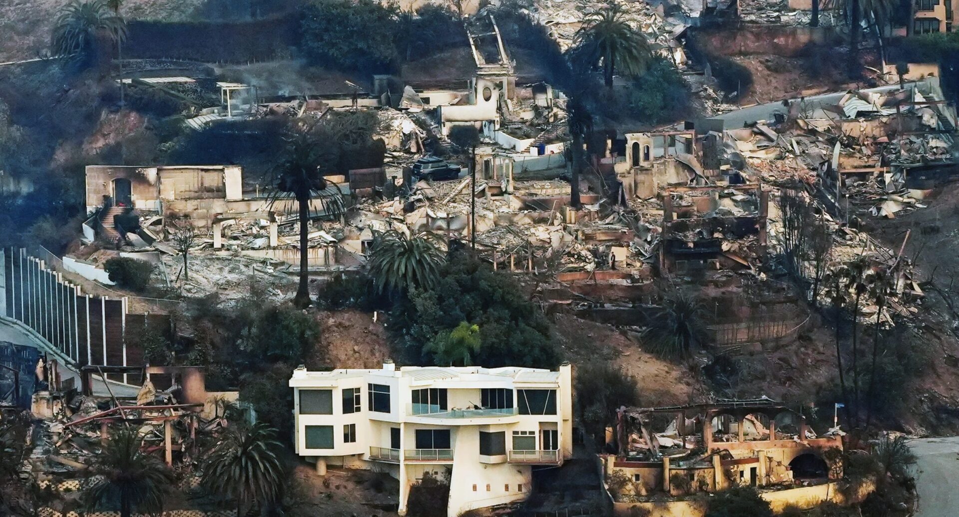 Aerial view of a hillside neighborhood where many houses have been reduced to rubble and ashes after a wildfire. One white house remains standing amid the devastation, surrounded by charred trees and debris.